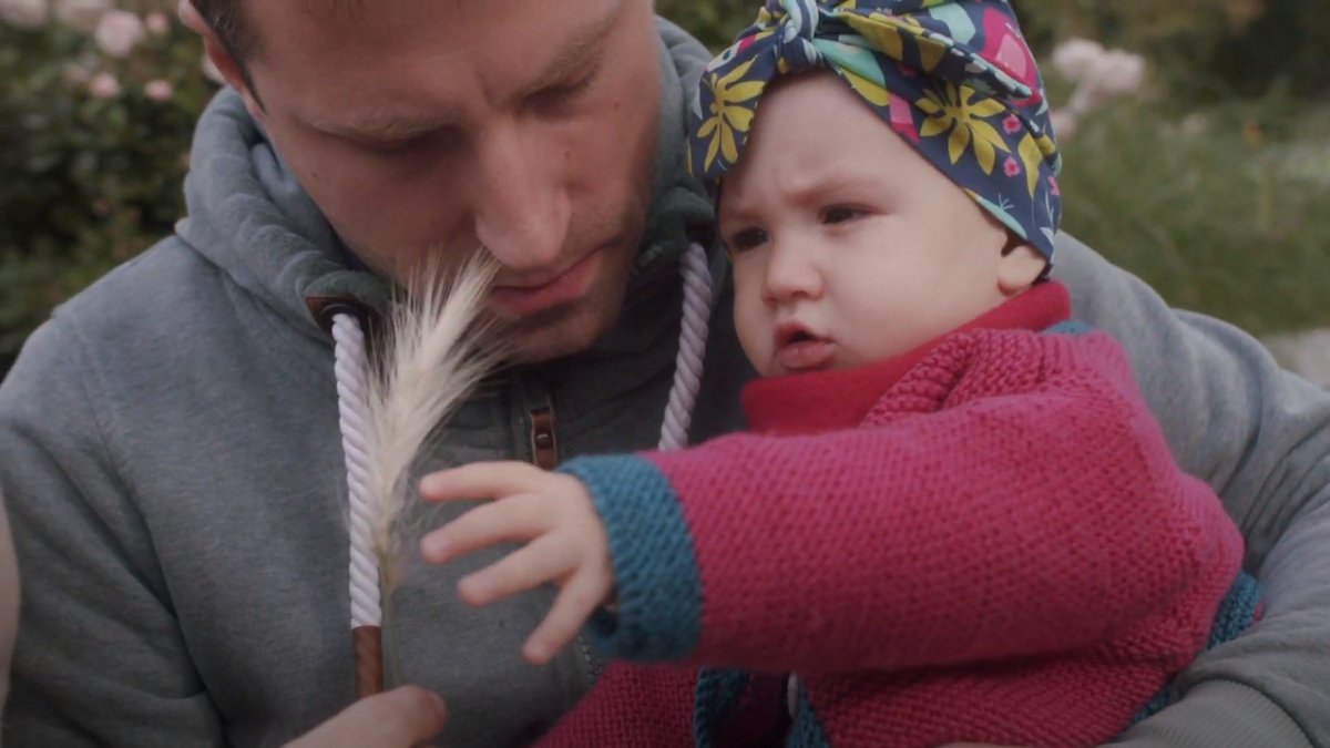 VBV Ihre Vorsorge im grünen Bereich Thumbnail Ein Mann hält ein Baby im Arm. Beide betrachten neugierig eine Ähre, die der Mann in der Hand hält. Das Baby trägt eine bunte Kopfbedeckung und eine pinke Strickjacke, der Mann einen grauen Hoodie