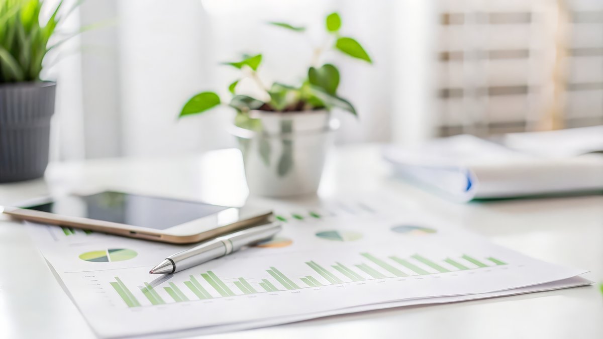 Bericht mit Tablet und Pflanzen Das Bild zeigt einen Tisch auf dem Zettel, ein Tablet und ein Stift liegen. Im Hintergrund sind zwei Pflanzen am Tisch zu sehen.