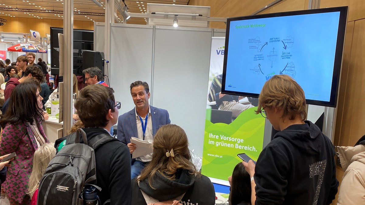 VBV Schülermesse 2024 Messestand der VBV-Vorsorgekasse auf einer Veranstaltung. Ein Mitarbeiter spricht vor dem Stand zu einer Gruppe junger Besucher:innen. Über dem Stand ist das VBV-Logo mit dem Slogan „Ihre Vorsorge im grünen Bereich“ sichtbar. Rechts zeigt ein Bildschirm eine Grafik zum Thema „Systematik Meldewege“. Der Stand ist gut besucht, im Hintergrund sind weitere Aussteller und Besucher:innen zu sehen.