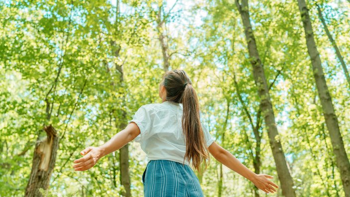 Junge Frau steht im Wald Das Bild zeigt eine junge Frau, die im Wald steht und die Arme hebt.