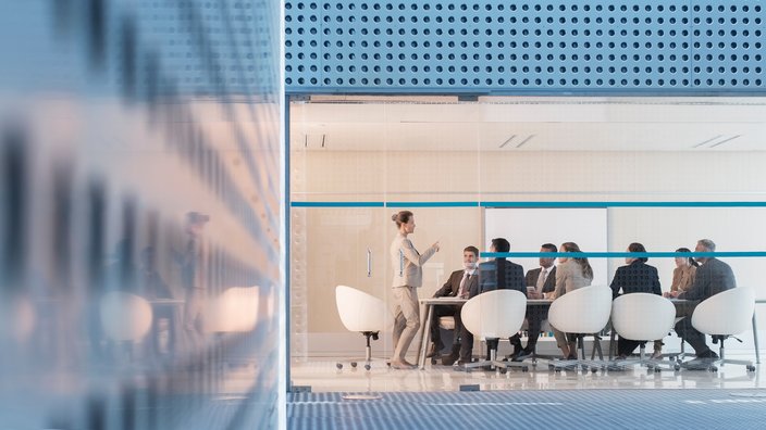 Besprechung im modernen Konferenzraum Ein moderner Konferenzraum mit Glaswänden, in dem ein Geschäftstreffen stattfindet. Acht Personen sitzen um einen Tisch, und eine stehende Person präsentiert etwas vor der Gruppe. Der Raum hat ein sauberes, professionelles Erscheinungsbild mit weißen Stühlen und einem blauen Streifen, der horizontal über die Glaswände verläuft.