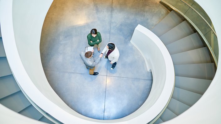 Business-Meeting im architektonischen Umfeld Das Bild zeigt drei Personen stehen in einem kreisförmigen Bereich am unteren Ende einer Wendeltreppe. Der Boden ist aus glattem, poliertem Beton und die Treppe hat ein modernes Design mit einer weißen Außenwand und grauen Stufen. Die Personen scheinen in ein Gespräch vertieft zu sein, wobei eine Person ein Klemmbrett oder Dokument hält.