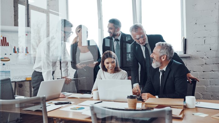 Professionelles Teamwork im Konferenzraum Das Bild zeigt eine Besprechungssituation von sechs Personen, davon zwei Personen sitzend und vier Personen stehend in einer entspannten Atmosphäre mit Blick auf einen auf dem Tisch stehenden Laptop.