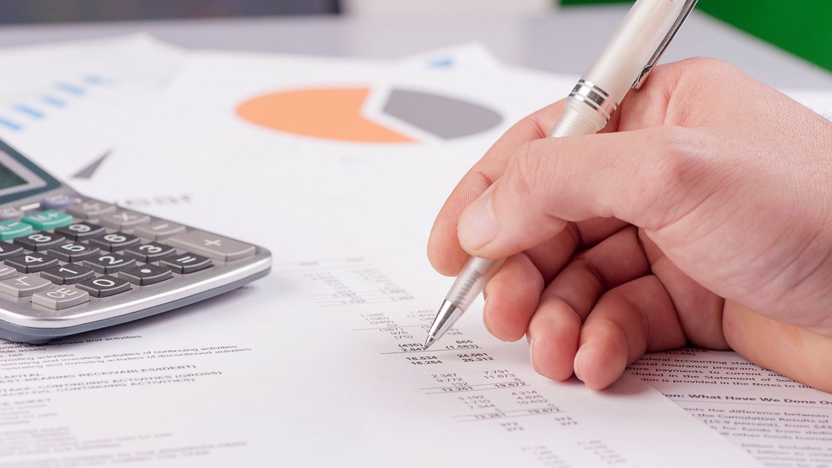 Afrac Stellungnahme 2016 Hand mit Kugelschreiber zeigt auf eine Finanzübersicht mit Zahlen und Tabellen; daneben liegt ein Taschenrechner, im Hintergrund ein Kreisdiagramm auf einem weiteren Blatt Papier.